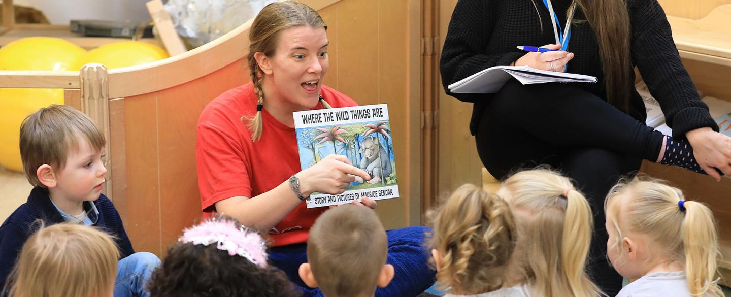 A woman in a red T-shirt reads to a group of children Where The Wild Things Are