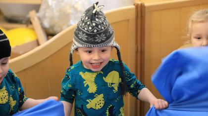 A young child smiling wearing a wooly hat and a green jumper with yellow and black dinosaurs