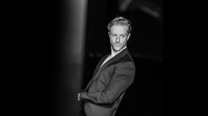 Black and white photo of blond man wearing a jacket sits leaning back looking over his shoulder.