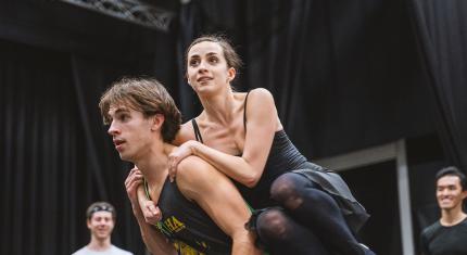 A dancer being carried on the back of another, both with determined looks