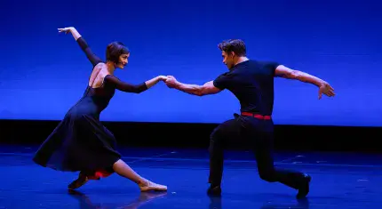 Against a blue back, two people in black dance holding one hand