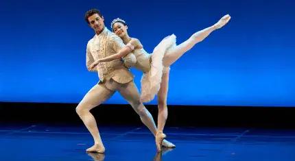 Two ballet dancers, a man and woman, stand next to each other, the woman on one leg leaning against the man