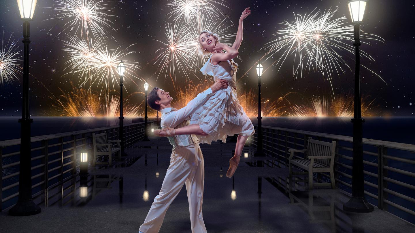 Male dancer holds female dancer aloft on a pier with fireworks in the sky