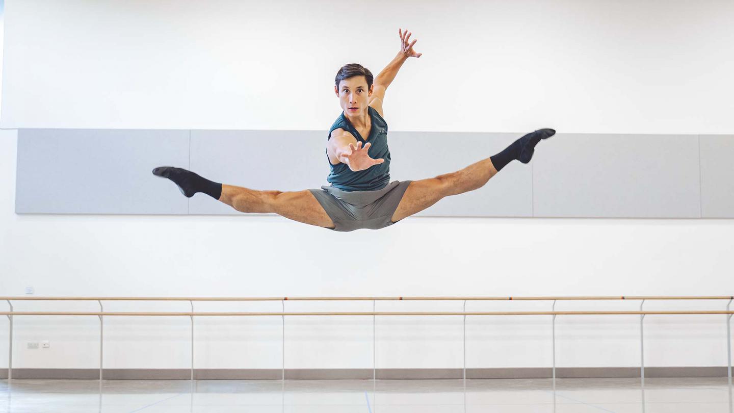 A male dancer jump with both legs extended in a mid-air split while reaching for the camara