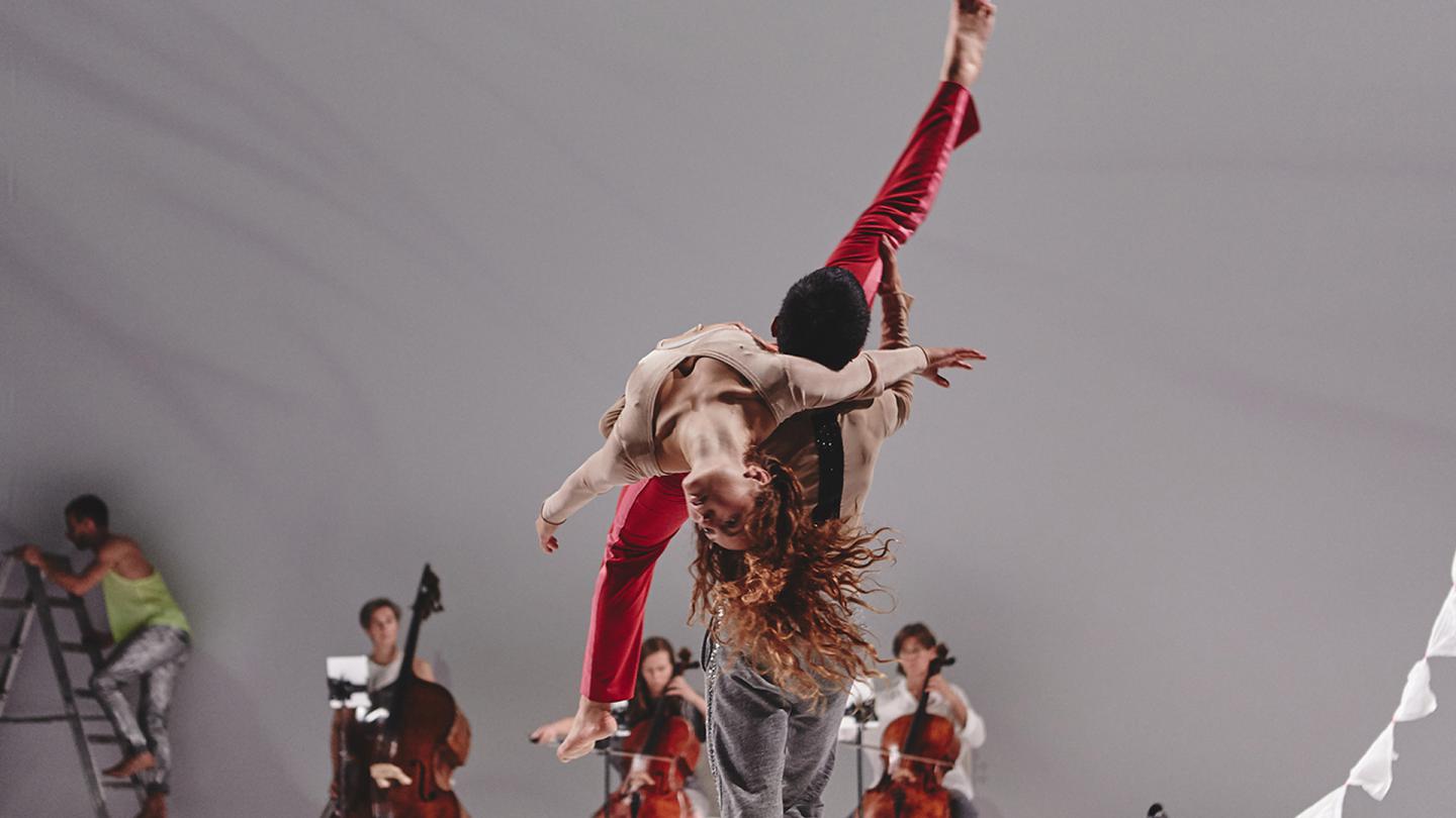 a dancer facing away from the camera holds a person wearing vivid red trousers on his shoulders, in the background a man climbs a stepladder and three people are sat playing their large stringed instruments