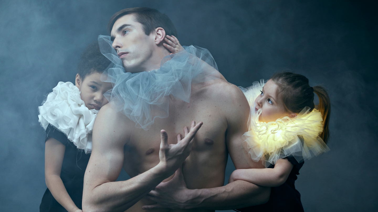 A man and two young children wearing colourful toule around their necks. They are surrounded by smoke with a dark grey background behind them.