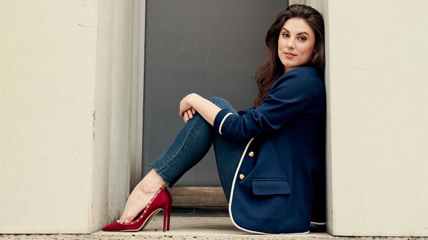Woman with long dark hair sat in jeans and a blue blazer wearing red shoes.
