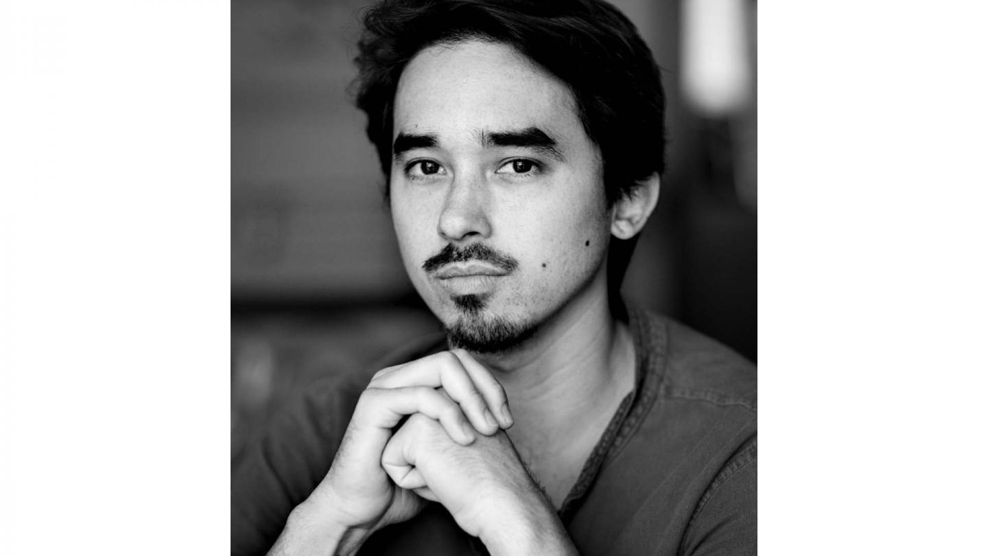 Black and white photo of man with facial hair and short dark hair looking directly at the camera, his folded hands in front of his chin