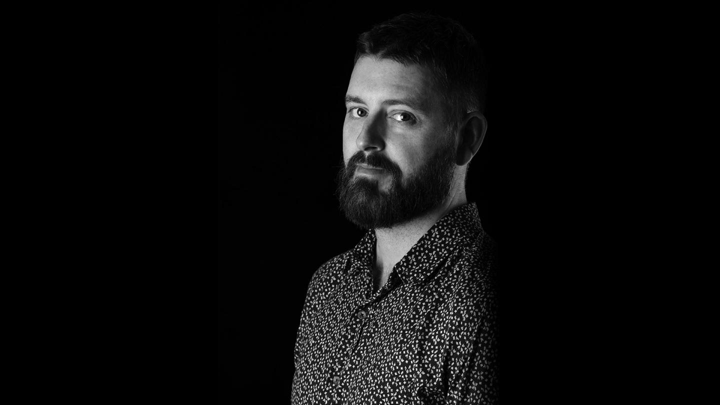 Black and whiote photo of Ben Wright in a patterned shirt, he has a beard, pale skin and dark hair.