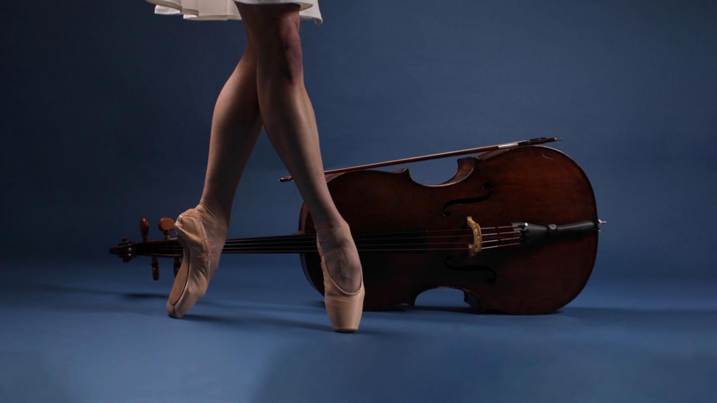Photo of the bare lower legs wearing pointe shoes in front of a viola places on its side all against a blue background