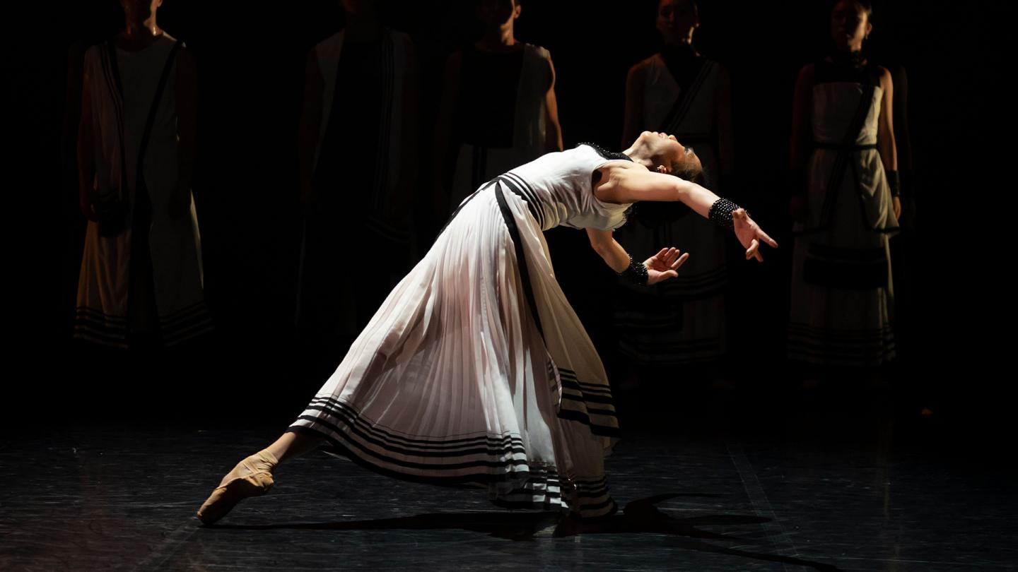 Dancer in a long white dress with black trim leans back, her torso level with her waist.