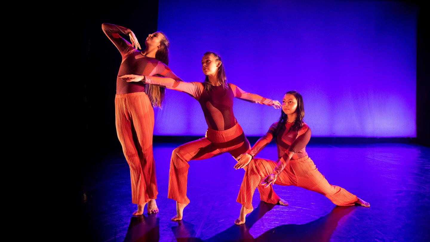 Three dancers bathed in red light against a background of black and blue, each in a different pose - one on her toes sood tall, the next bending one leg at 90 degrees, the third lower still