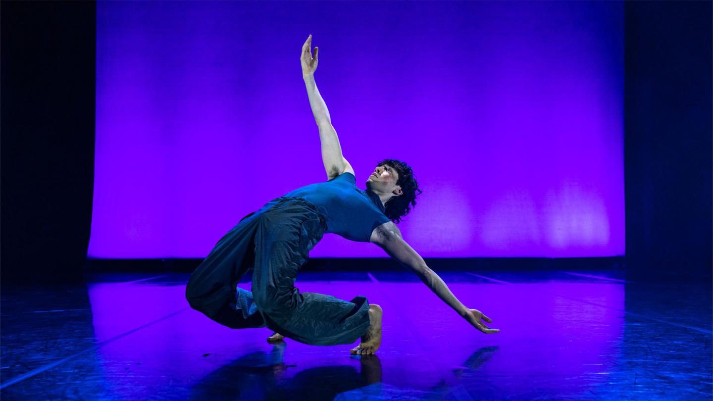 male dancer on dark trousers and a tight blue top bends backwards, nearly on the ground but still balanced despite being bent double backwards.