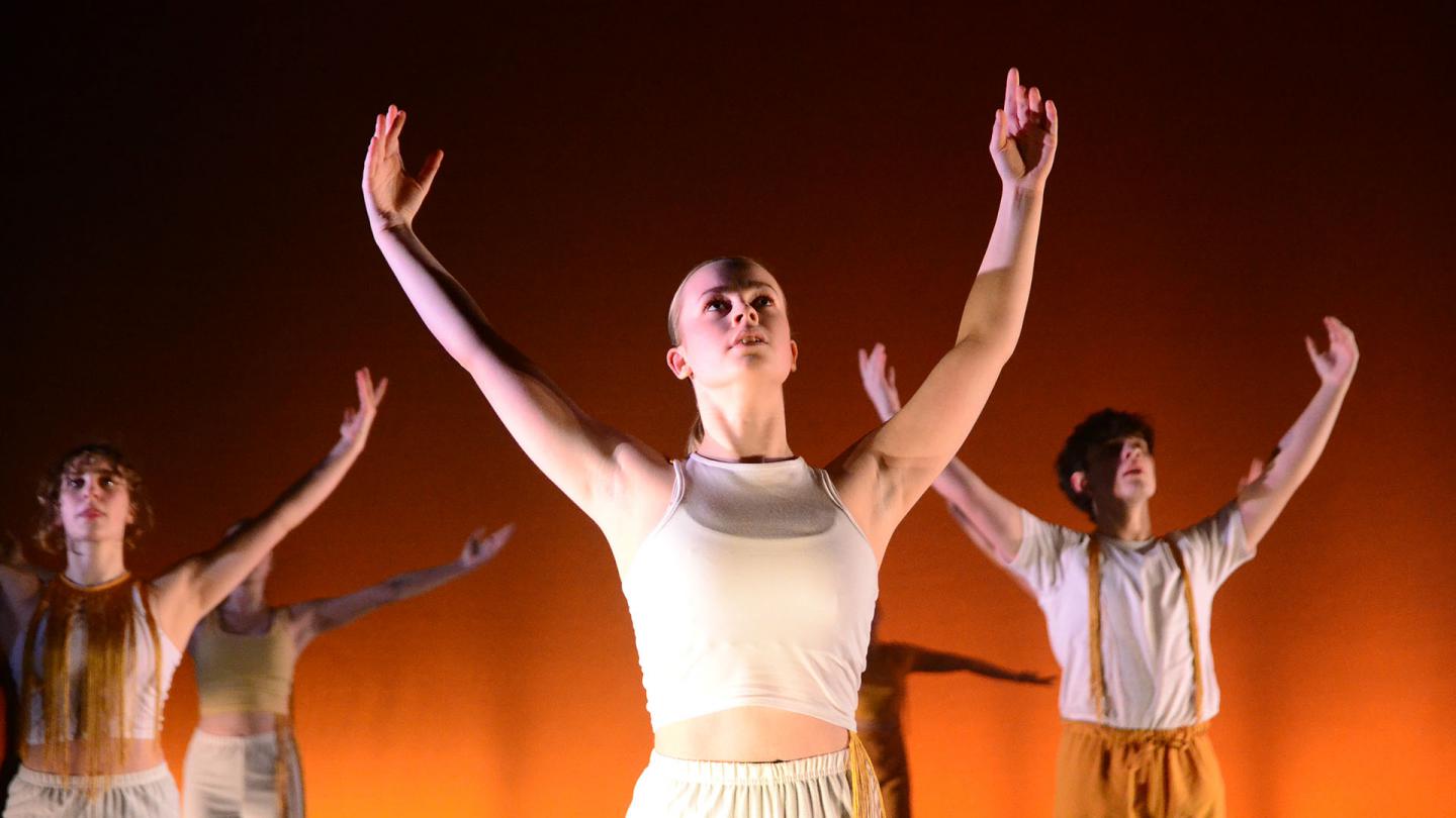 Young female dancer in a white top against a lit orange background looks into the air and raises her hands above her head