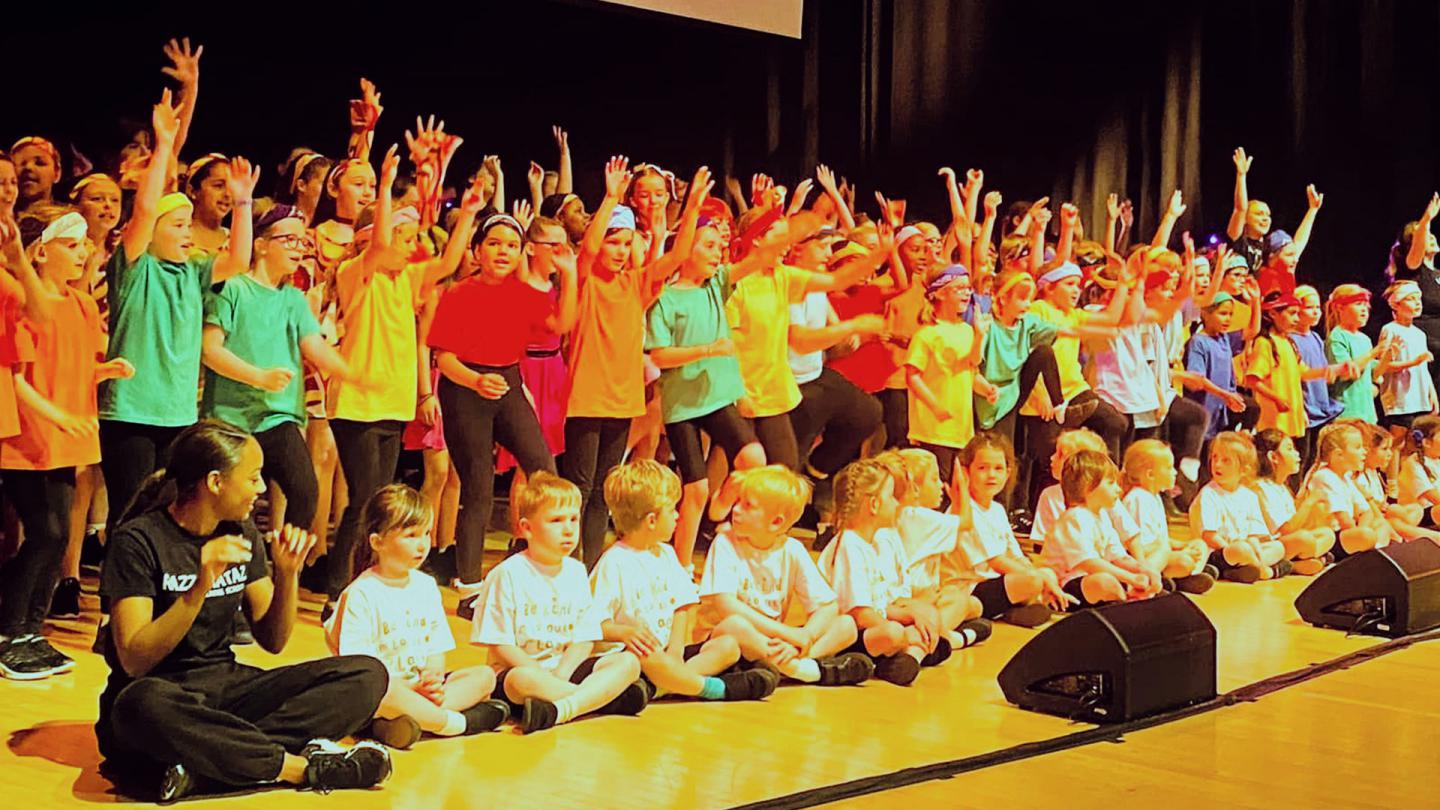 Group of over 50 children wearing different colours all waving out from a stage happily