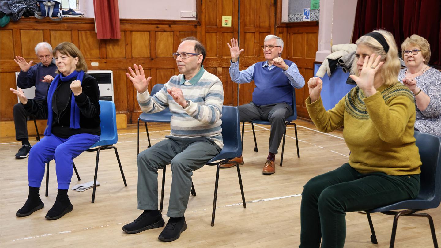 A group of people sat in plastic chairs raising their hands copying an unseen instructor