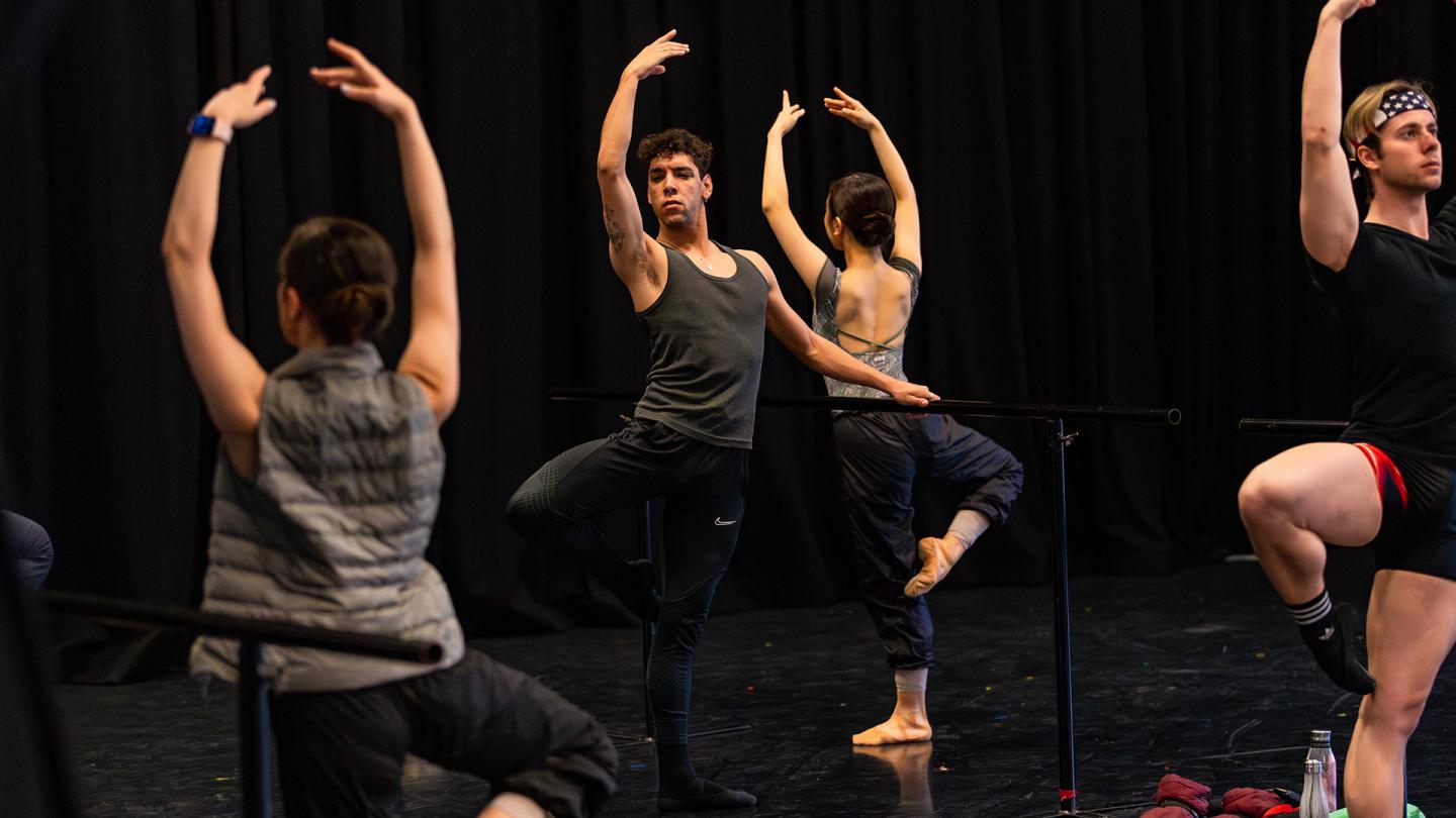 Dancer in a dark vest, one hand hovering above a barre, the over raised above his head, standing on one foot with the other leg raised at the knee