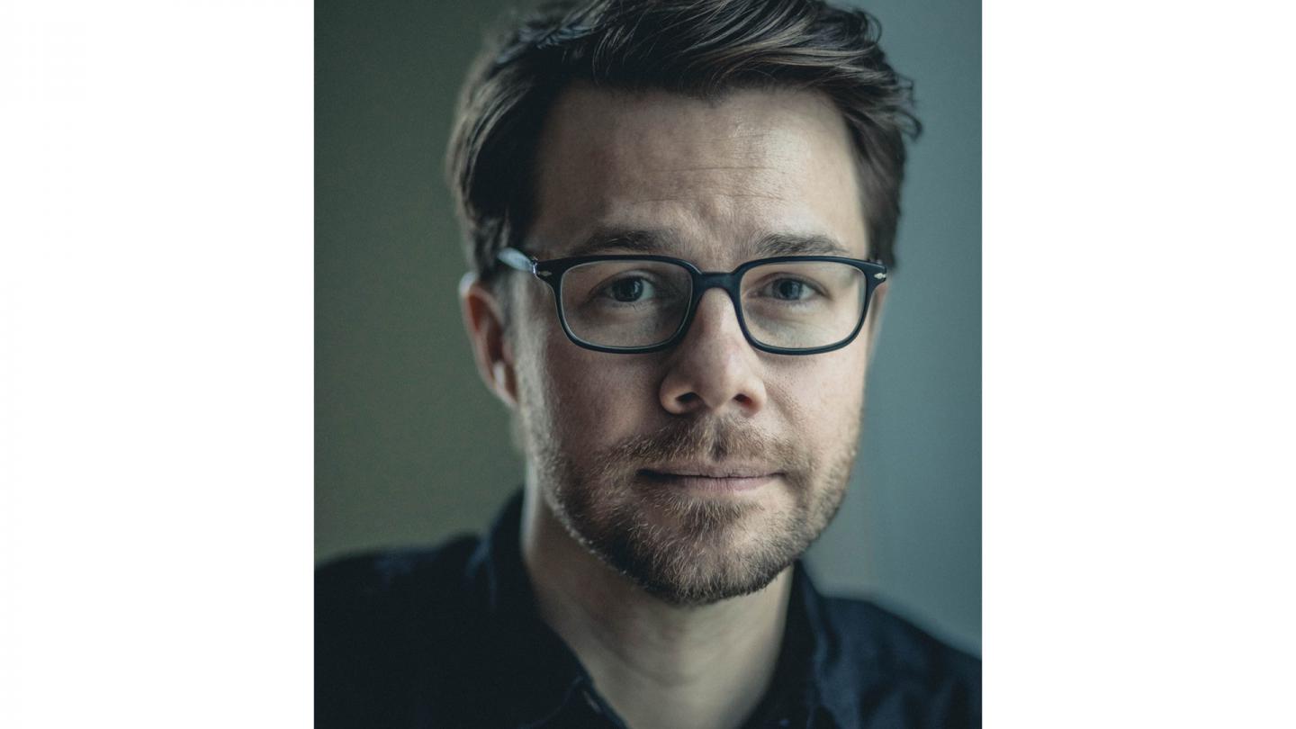 White man with brown hair, a beard and glasses wearing a shirt with the neck open