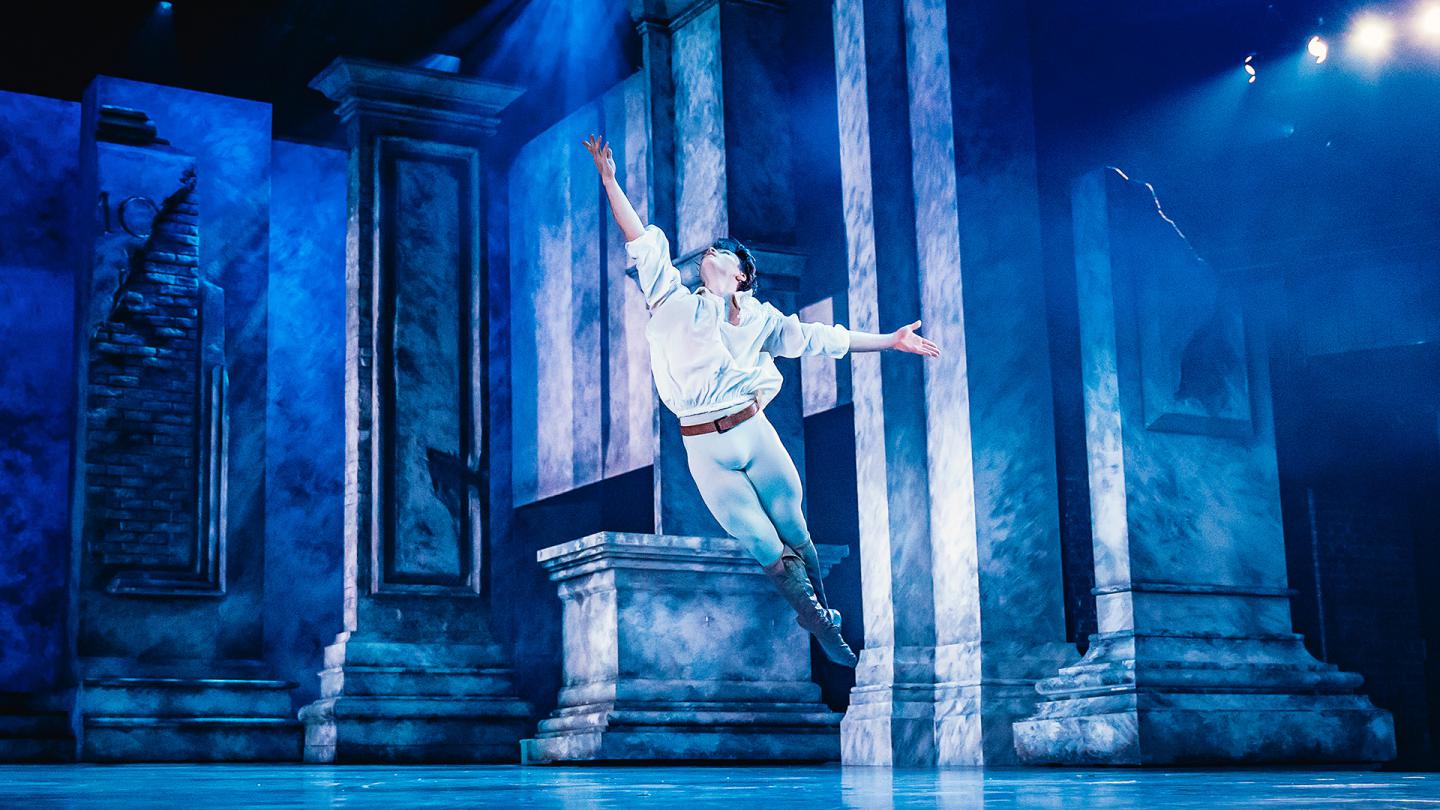 Dancer leaping across the stage, his legs together, one arm raise triumphantly above him, bathed in blue light