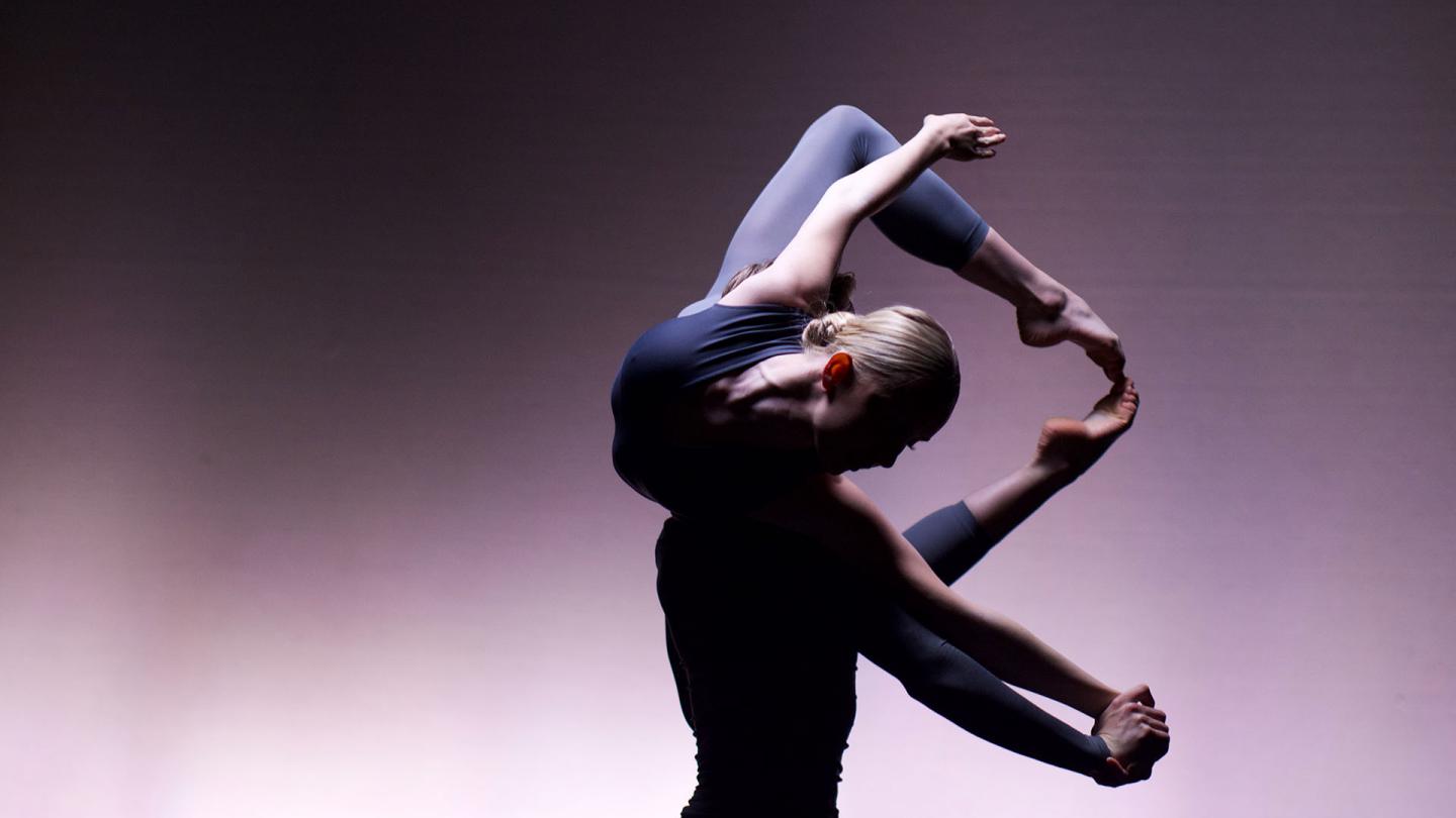 Against a gradiated purple backdrop one dancer lifts another, the lifted dancer horizontal, toes together but knees apart back curled around the lifters head