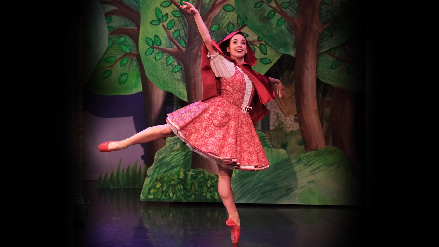 Dancer in red with a red hood and red pointe shoes stands arabesque smiling as she walks through a forest