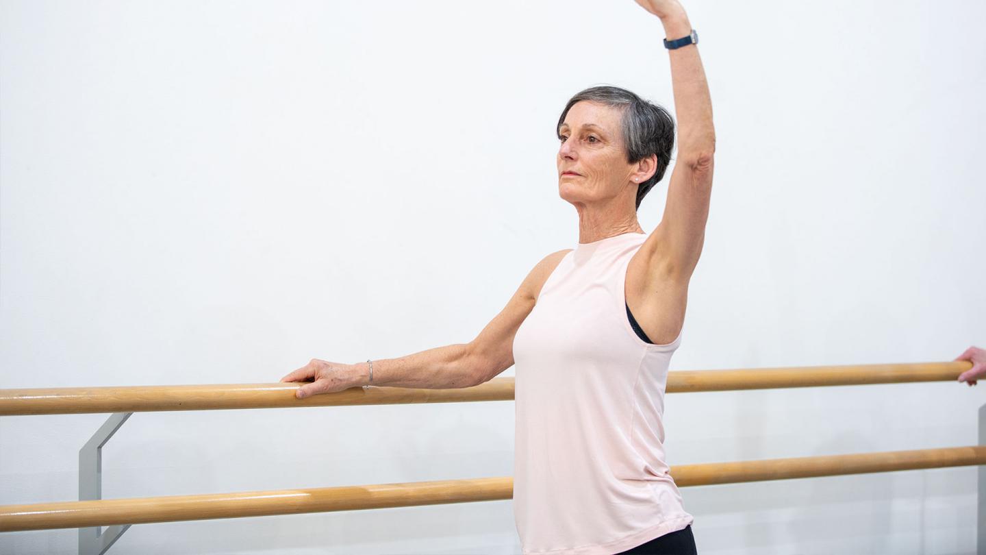 Standing at the barre with one hand holding on, a student of the Over 55s class wearing a pink top raises her other arm high while focusing in front of her