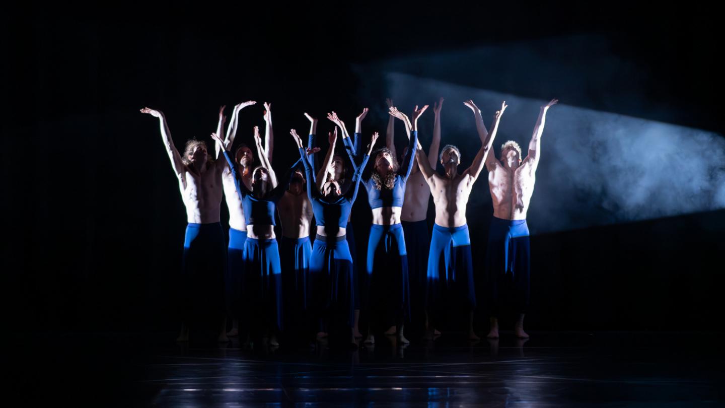 On a dark background, a group of dancers in blue outfits stand in a spotlight while reaching their arms up.
