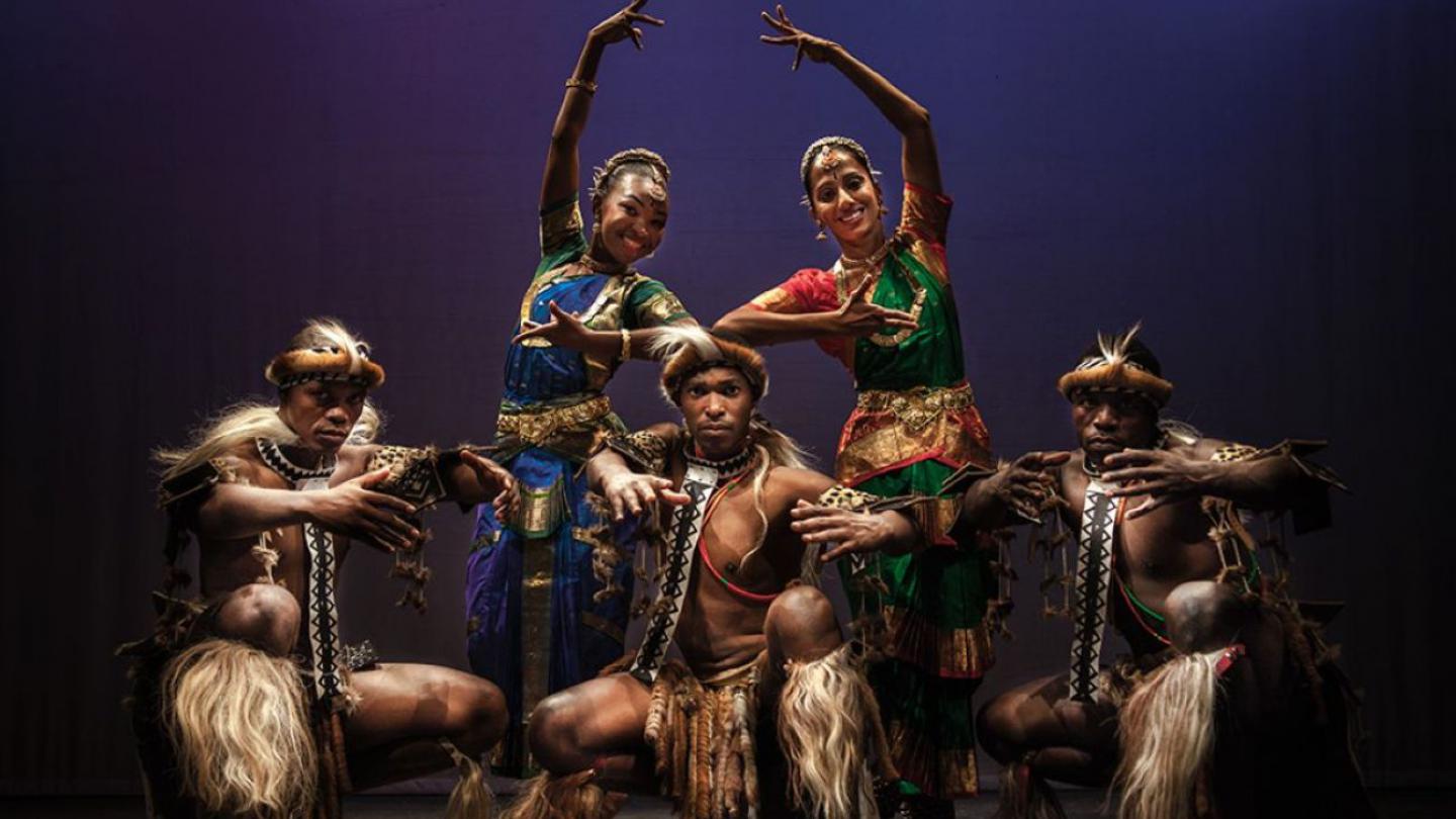 A group of five dancers in traditional costumes strike a pose. Two women are positioned in the centre wearing colourful attire and holding their arms elegantly over their heads. The men, adorned in earthy tones with fur-like accessories, crouch in front. 
