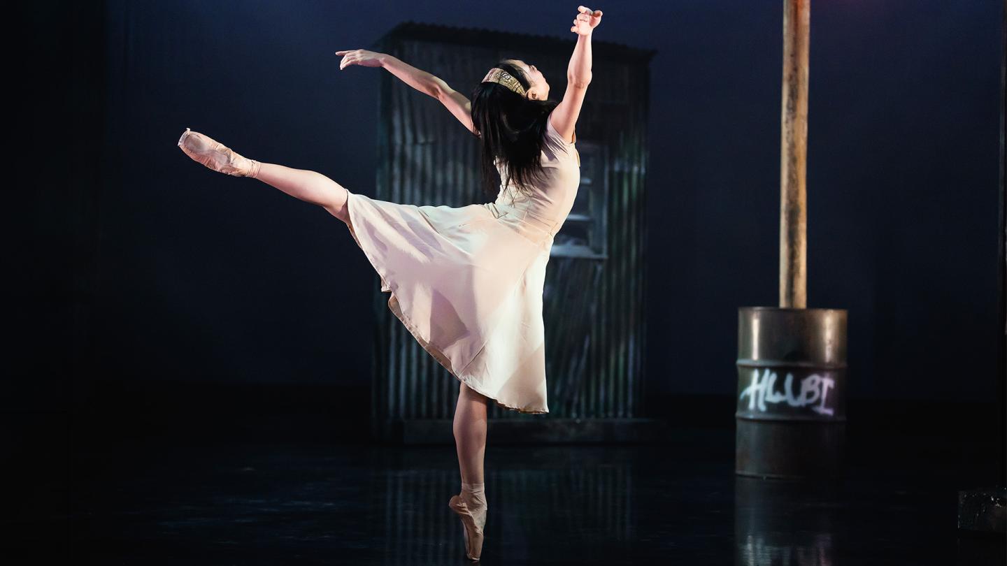 Dancer in a pale dress wearing a hair band stands on one foot en pointe, a leg straight behind her, both arms above her head