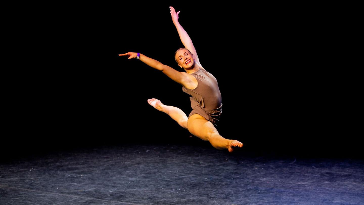Woman on a stage performing a grand jete wearing a brown top and shorts, slicked back hair and an open smile