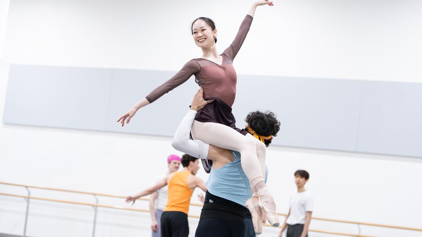 Dancer smiling as she is lifted, sat on the left shoulder of another dancer, one arm angled up, the other angled low