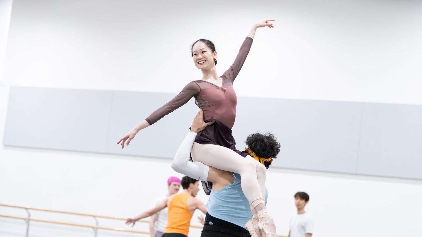 Dancer smiling as she is lifted, sat on the left shoulder of another dancer, one arm angled up, the other angled low