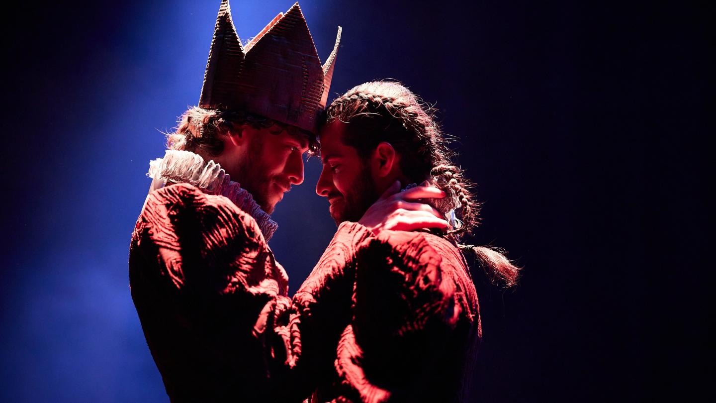 On a dark background, two male dancers are facing each other in an embrace. Their foreheads are touching and they are looking into each other's eyes. One of the men is wearing a large crown on his head.