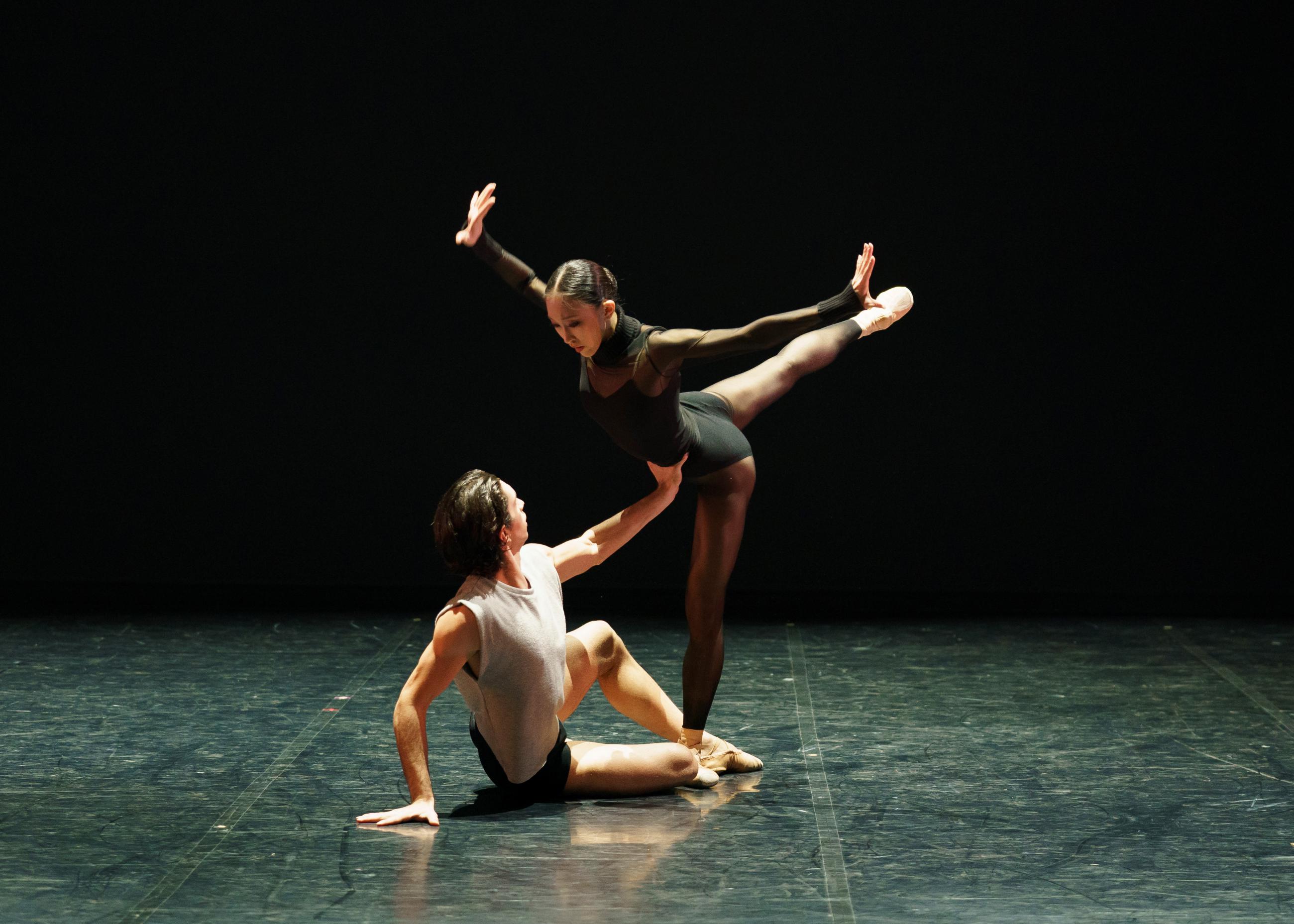Dancer Lorenzo Trossello sits on the floor supporting dancer Minju Kang in ballet position.