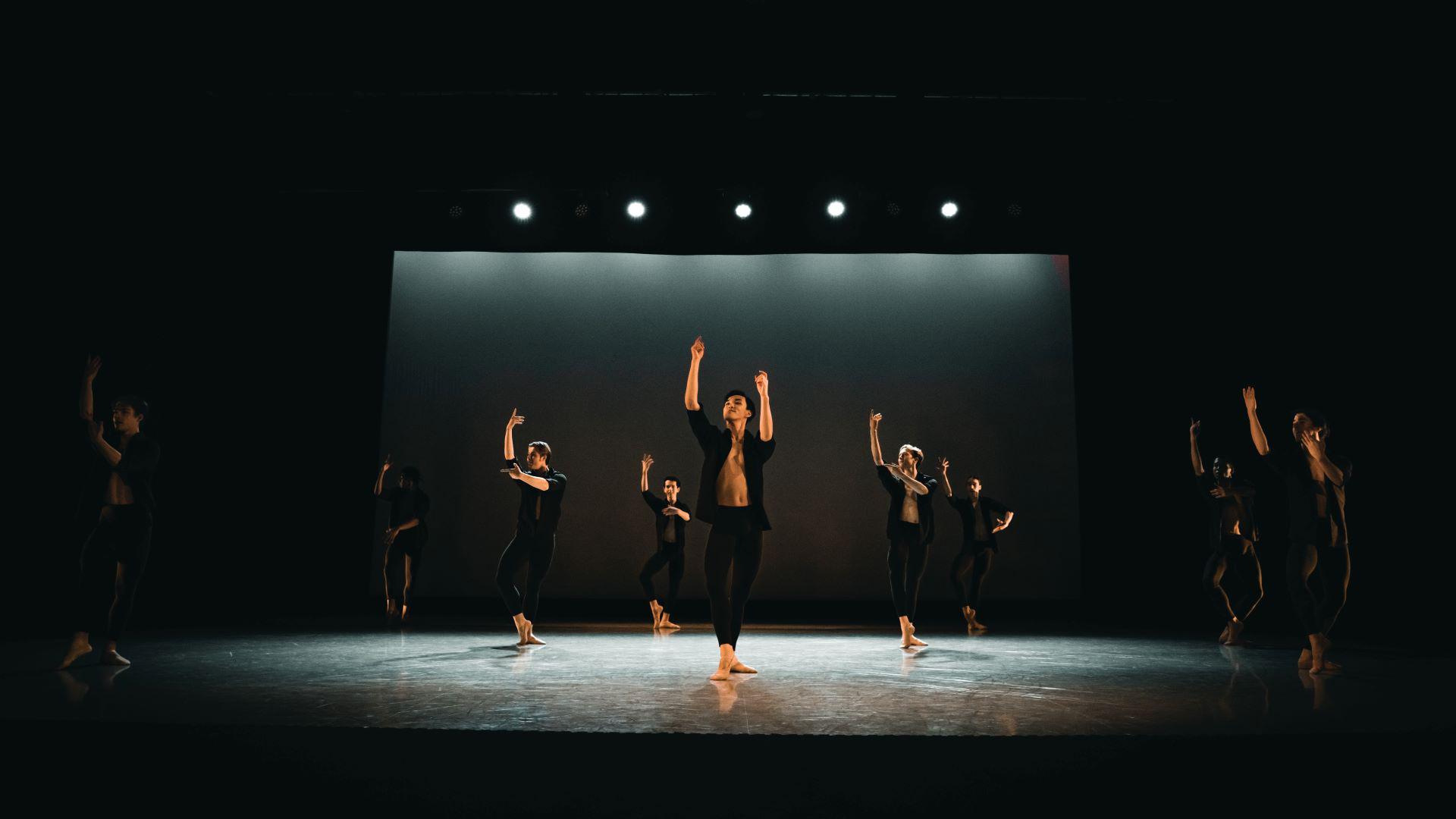 Male dancers in black trousers and open black shirts strike synchronised poses, pointing to the sky