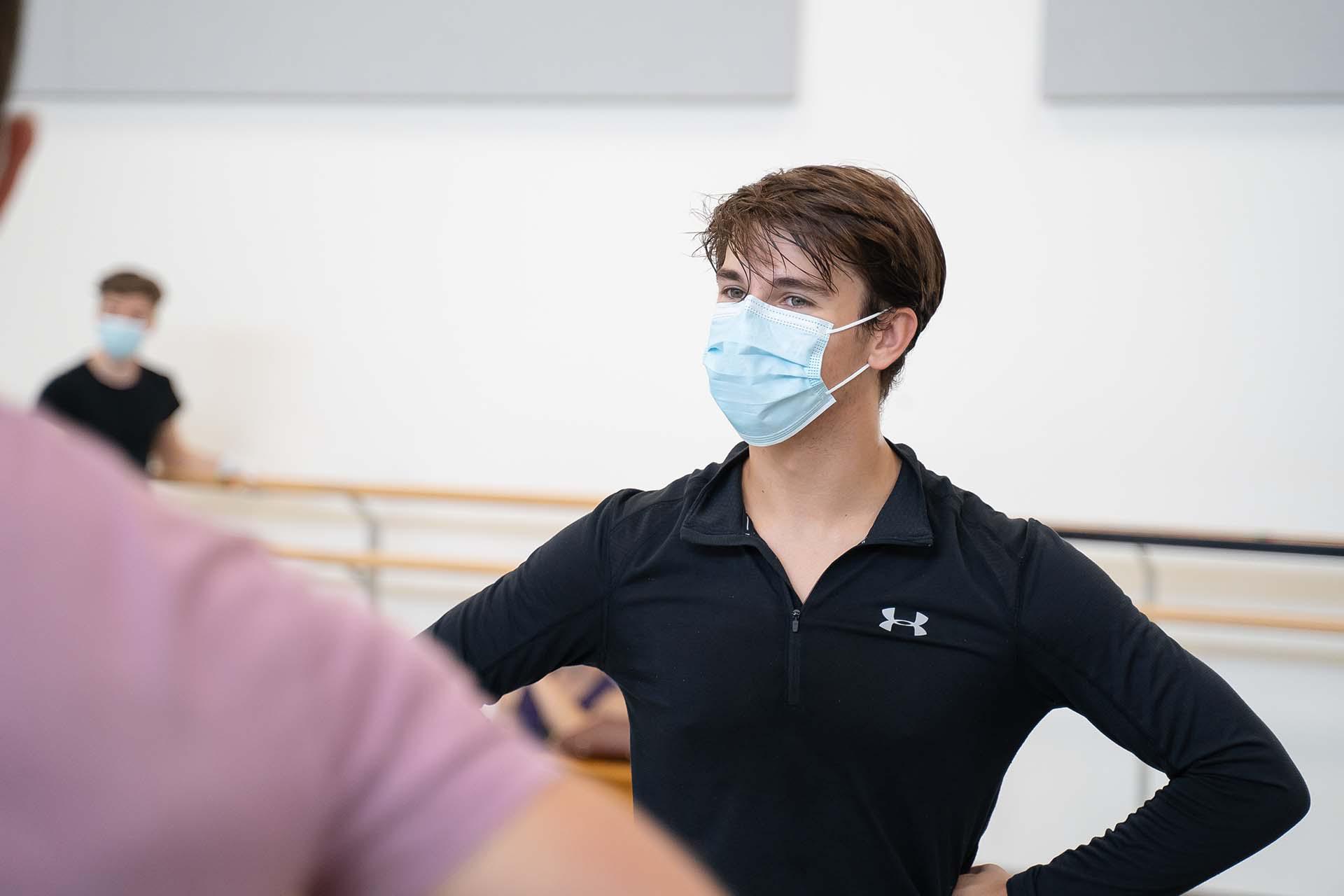 A dancer stands with hands on his hips while receiving instruction from the choreographer