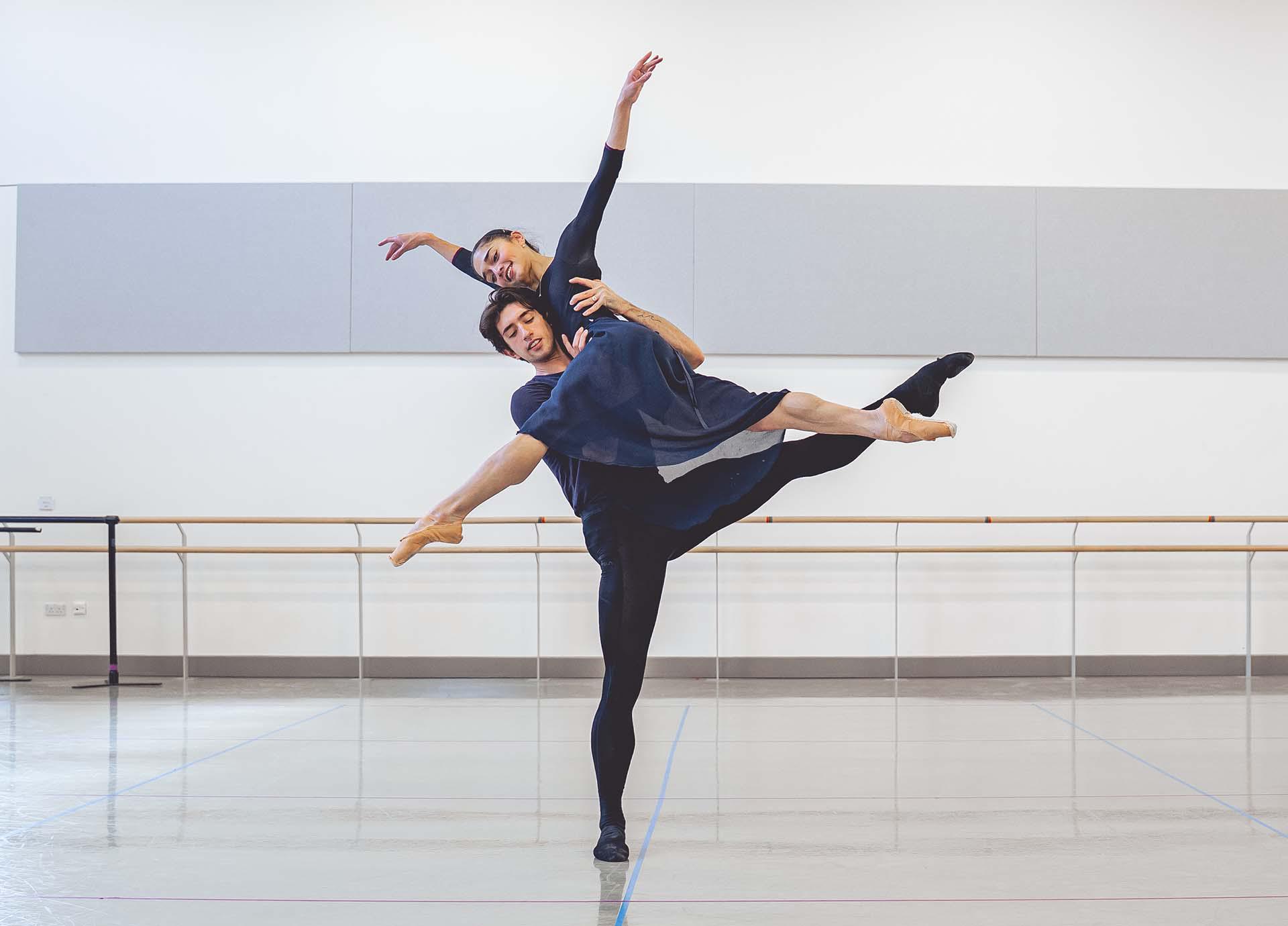 A male dancer hold a female dancer on his shoulder who she extends one leg in front of her and another behind