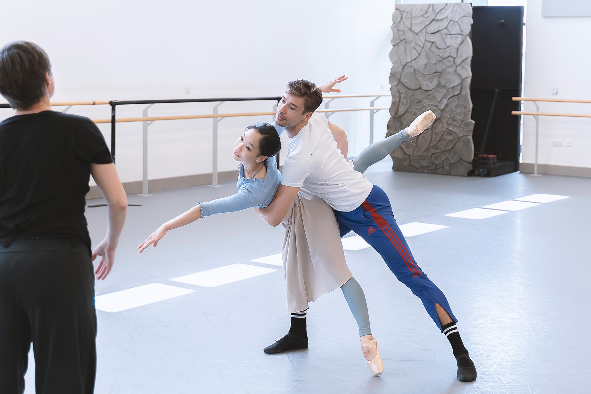The rehearsal director looks on as two dancers embrace in a difficult ballet pose