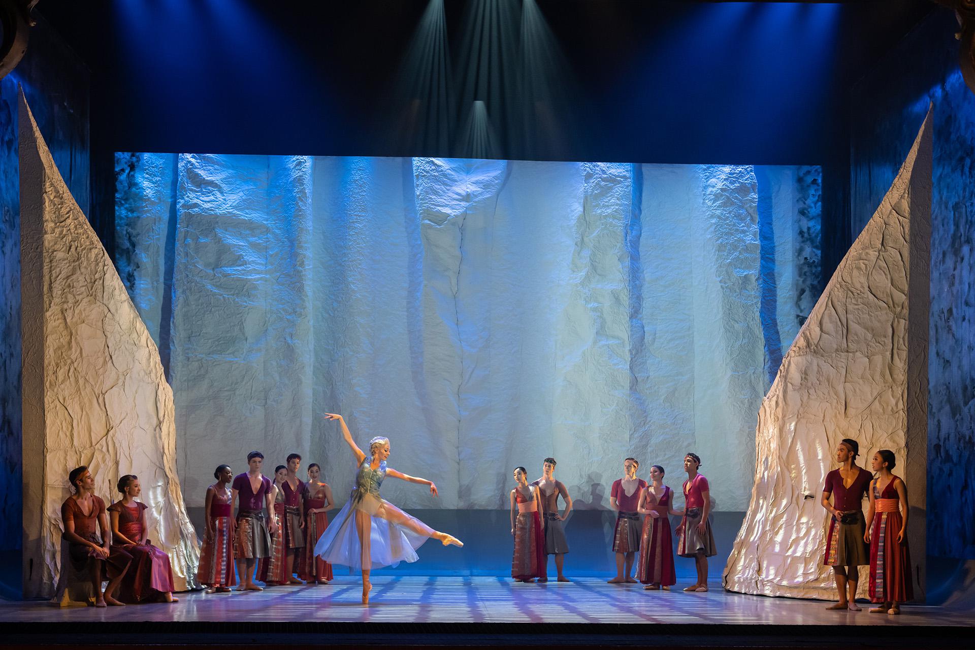 Marilla, in a light shiff and blue top, dances in front of the gathering on land, all wearing reds, and they are all watching her