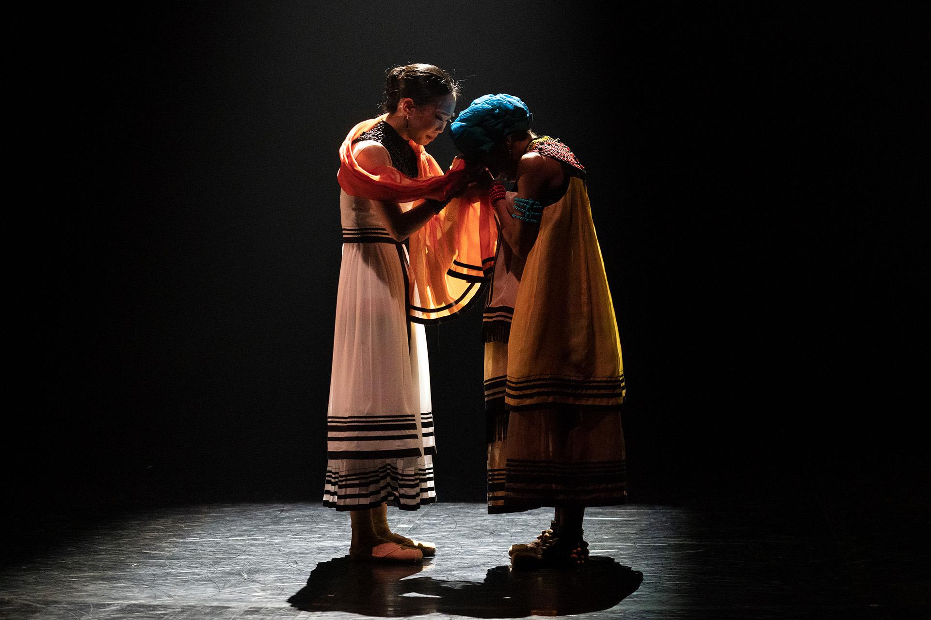 Mother and daughter stand in front of each other, heads bowed against a black background