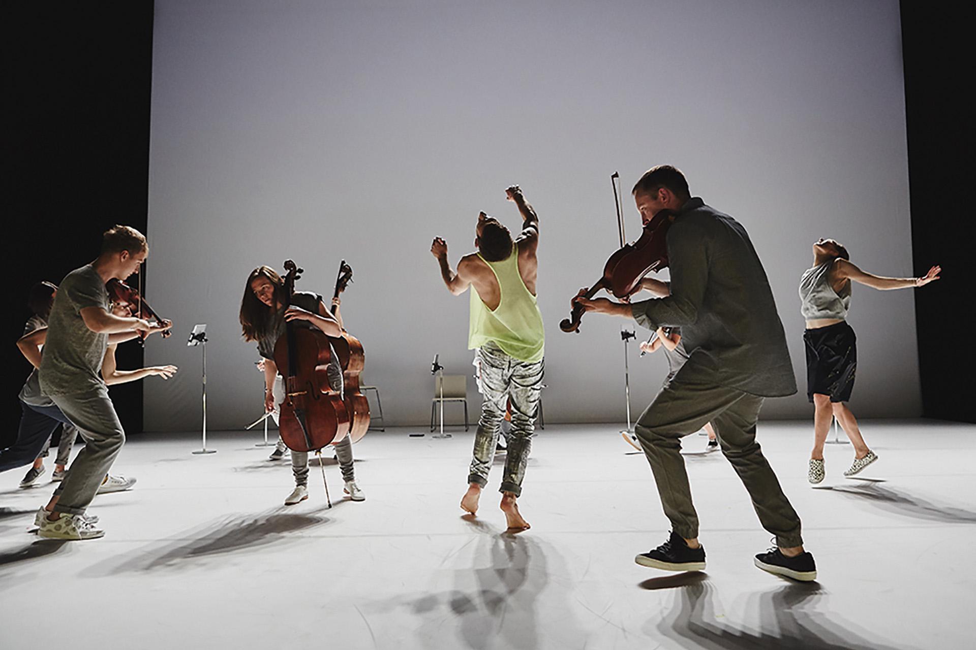 Violinists and a cellist in casual clothes play to a dancer in bare feet, shiny trousers and a yellow top