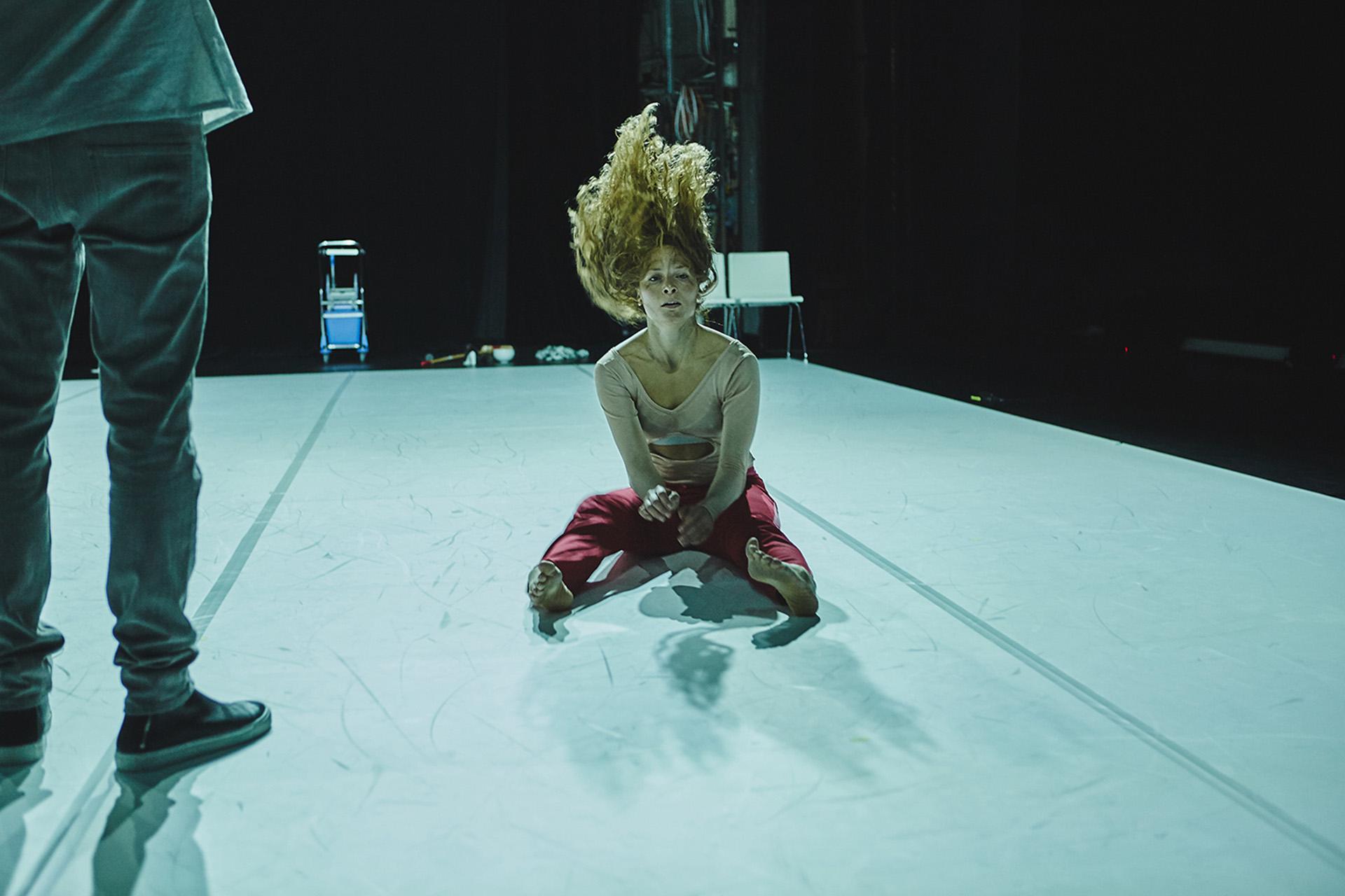 A persona in red trousers and a pale pink shirt sits on a white floor, her long hair is raised above her head as the photo is captured as she swings in back