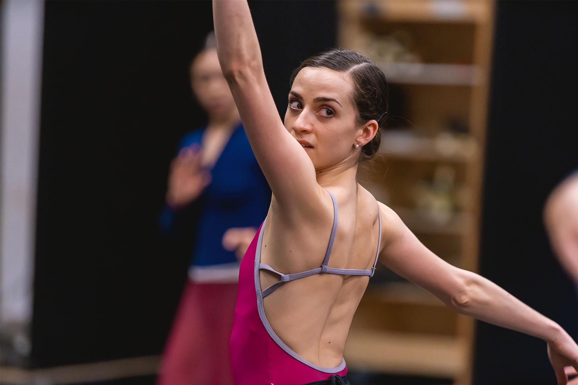 Woman dancer with dark hair tied in a bun looks back over her shoulder with one arm raised above her head