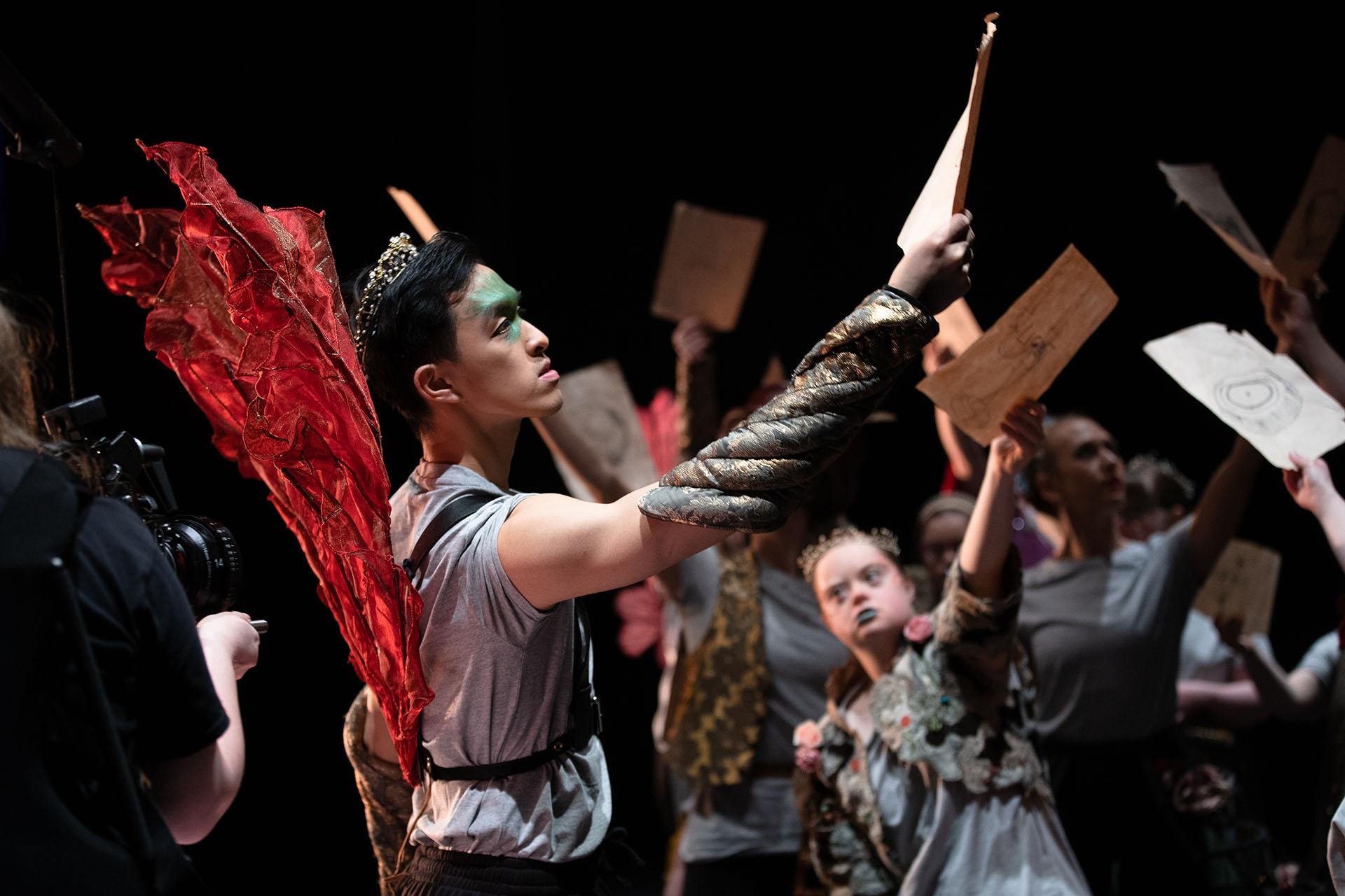 Dancer wearing red wing and green make-up on his face, hold a piece of paper in front of him. Other performers are in the background doing the same.