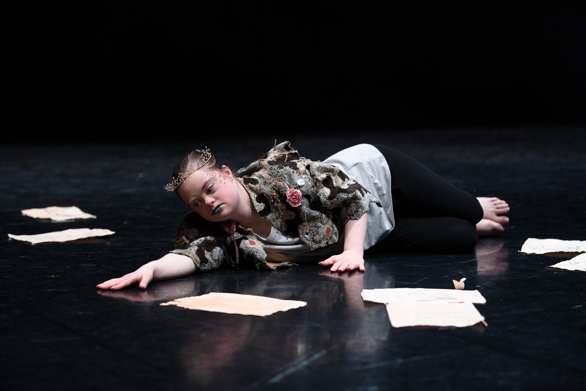 Performer laying on the black floor surrounded by discarded papers