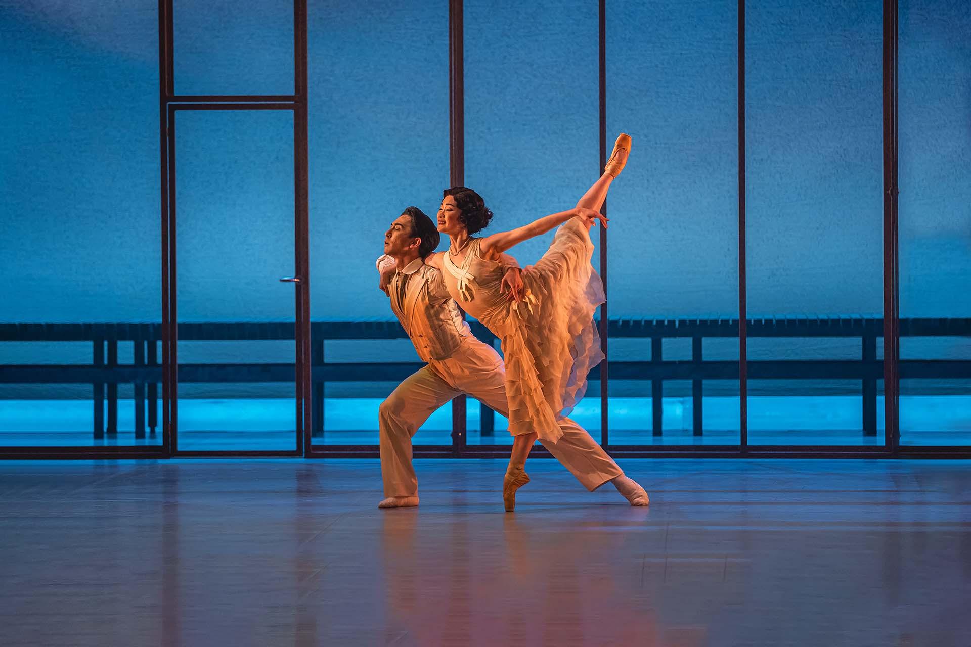 Two dancers in creamy white silk outfits dancer on a blue stage and look off stage lovingly