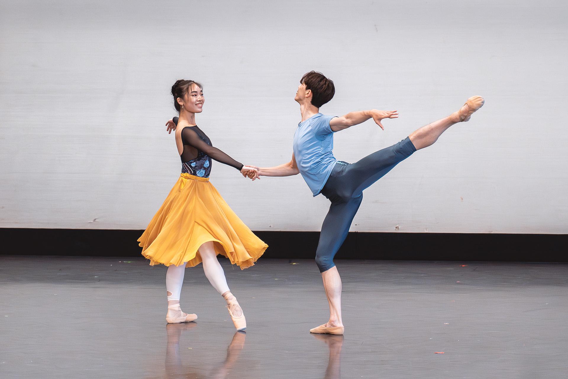 Two smiling dancers, one supporting the other with an outstretched hand, and the other, in a blue top and darker blue leggings, stands on one leg, his other stretched out behind him