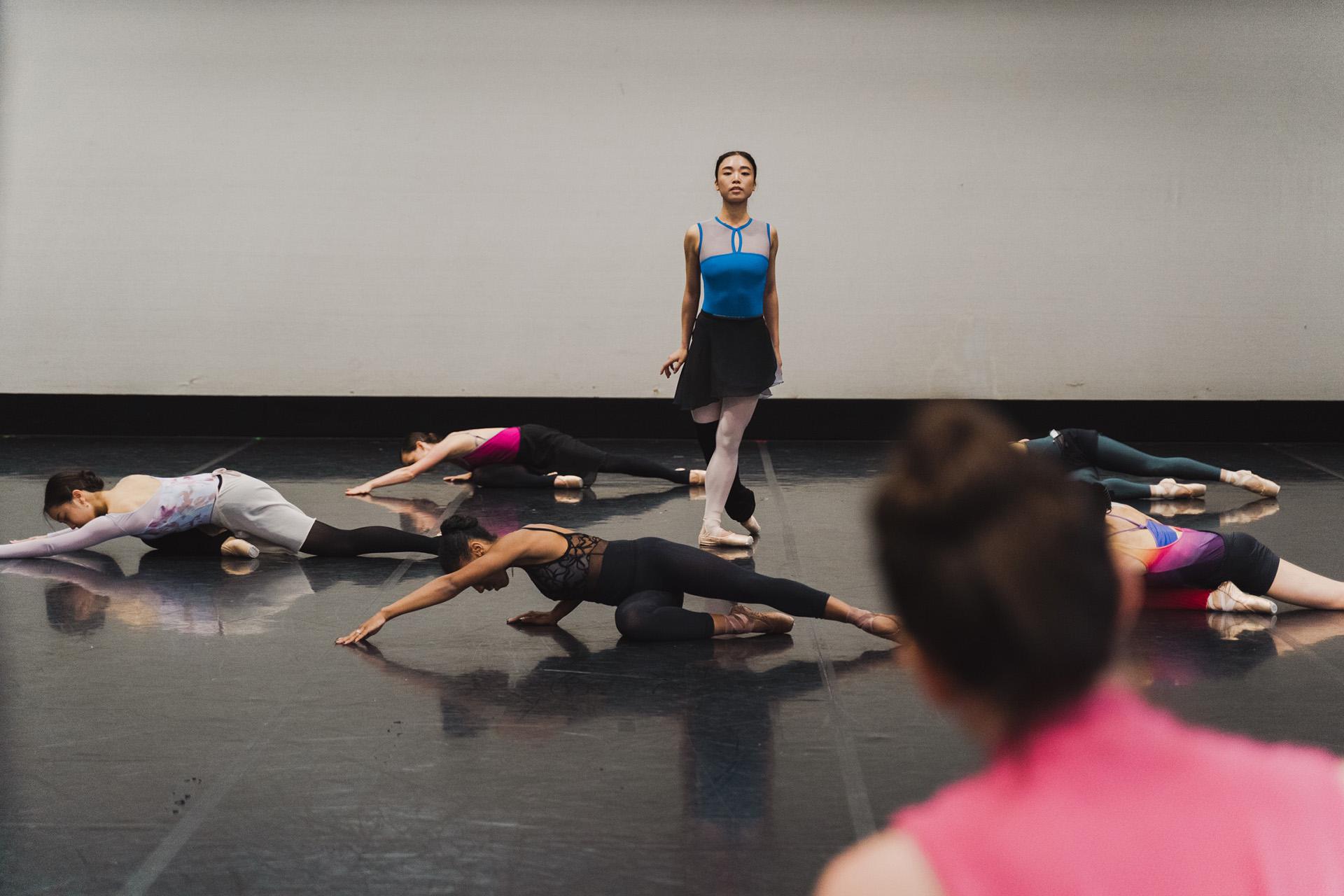 Choreographer looks on while a dancer walks imperiously while innumerate dancers surround her, all stretched across and floor facing down