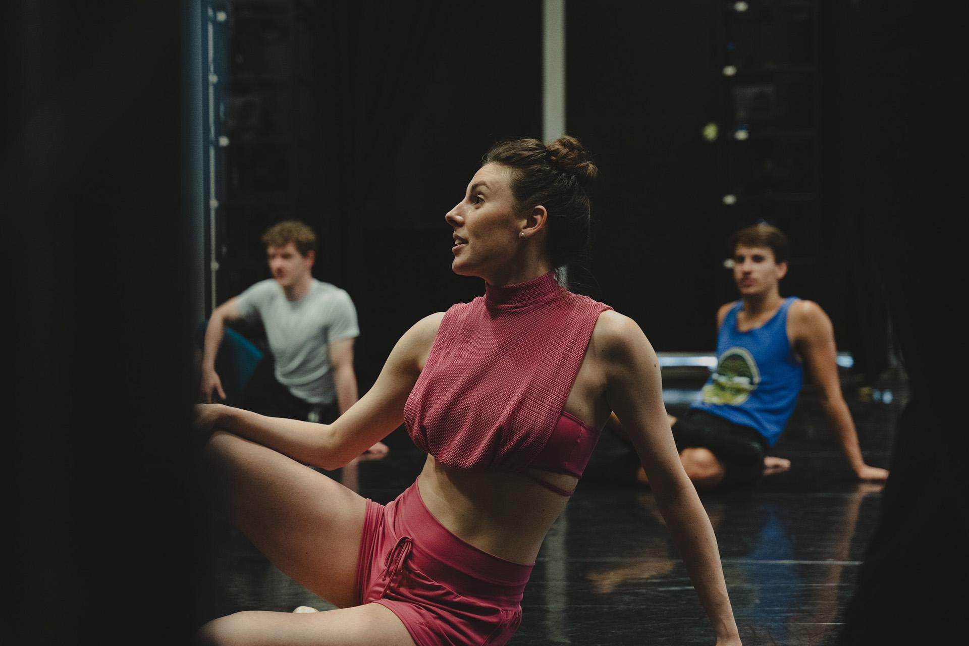Choreographer wearing pink, sat on the floor leaning back on one arm, while talking with someone out of frame