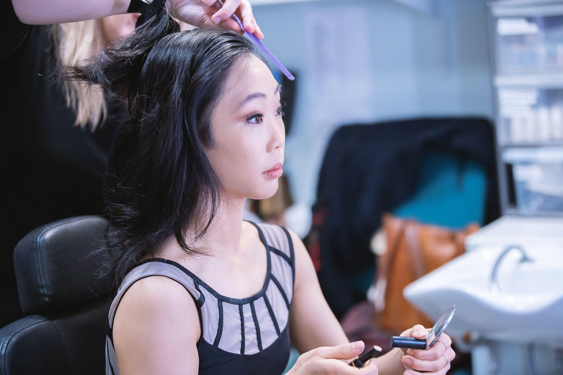 A female dancer sits as a hairstylist combs her hair.